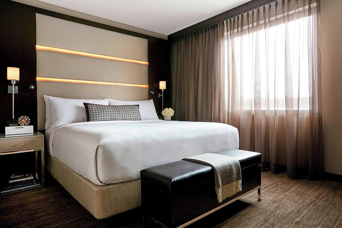 Ein modernes Hotelzimmer mit einem großen Bett, Nachttischlampen, einem Nachttisch, einer Bank und einem großen Fenster mit Vorhängen.