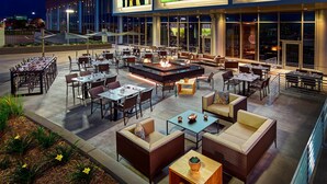 Un espacio de comedor al aire libre con mesas y sillas, un hogar para fogata y vistas de la ciudad por la noche.