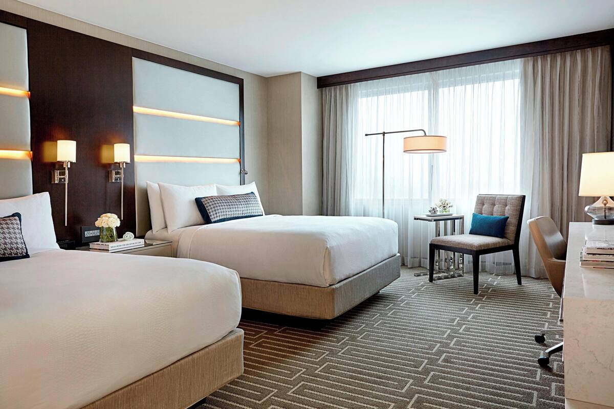 Ein Hotelzimmer mit zwei Betten, einem Schreibtisch, einem Stuhl und einem großen Fenster mit Vorhängen.