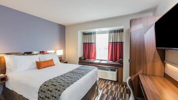 Habitación de hotel con una cama grande, una mesita de noche, un televisor, una ventana con cortinas y un banco empotrado.