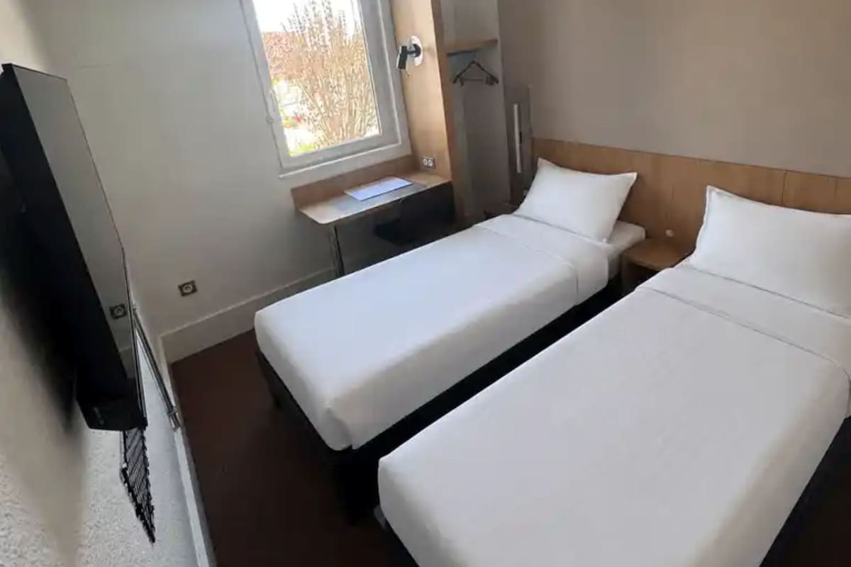 A hotel room with two beds, a desk, a TV, and a window.