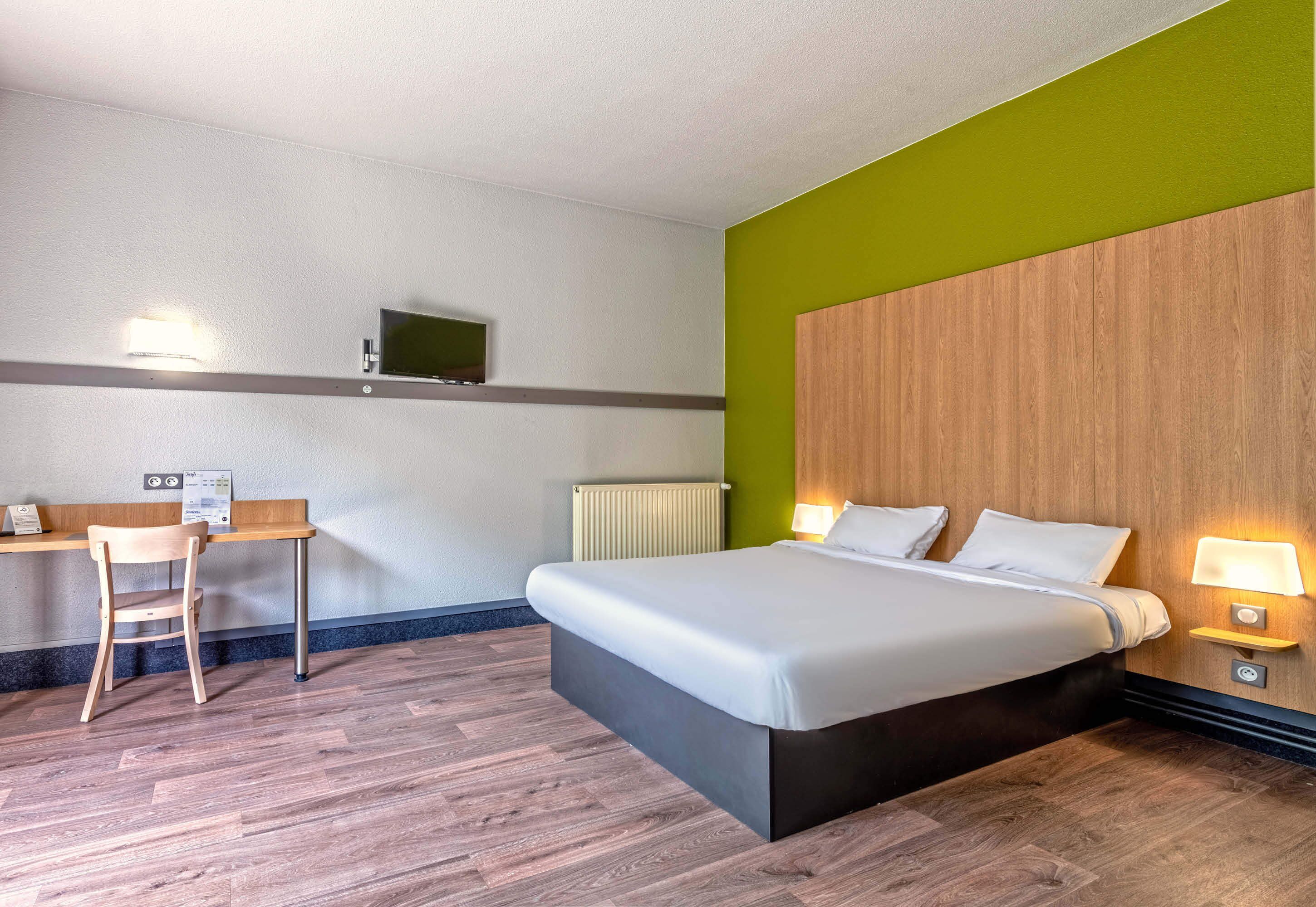 A hotel room with a bed, bedside tables, a desk, a chair, a television, and a radiator.
