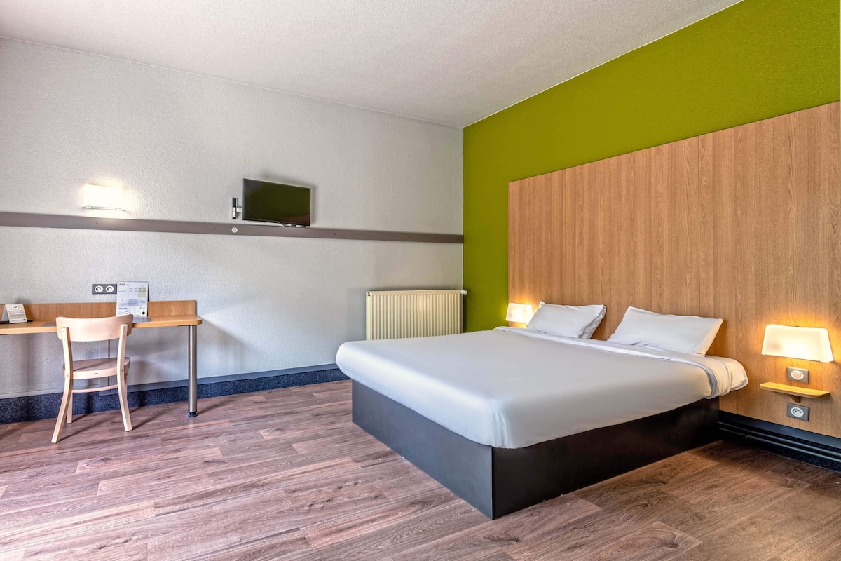 Quarto de hotel com cama, mesas de cabeceira, escrivaninha, cadeira, televisão e radiador.