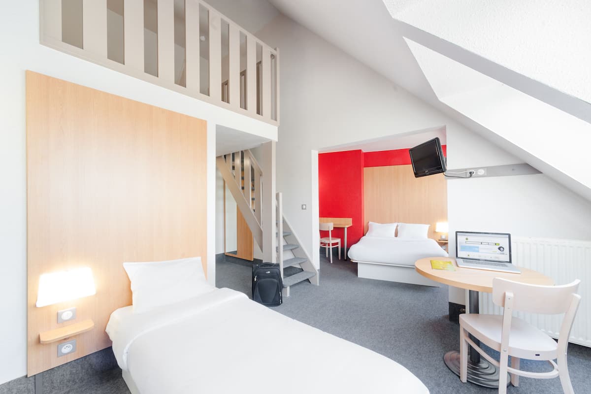 Quarto de hotel moderno com uma cama, uma mesa com um laptop, uma cadeira, uma escada e uma televisão na parede.