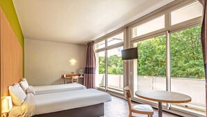 Habitación de hotel con cama, una mesita, una silla, televisor y ventanas amplias que ofrecen vistas al verdor exterior.