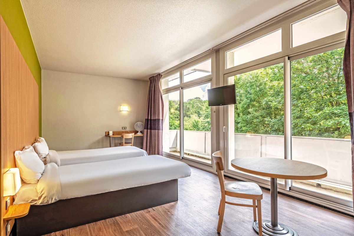 Quarto de hotel com uma cama, uma mesinha, uma cadeira, televisão e amplas janelas com vista para o verde lá fora.