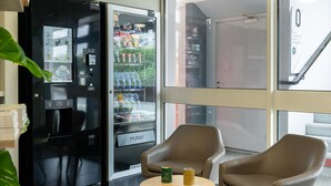 Máquina expendedora con un botón de 'PUSH', puerta de vidrio y una zona de estar con dos sillas y una pequeña mesa.