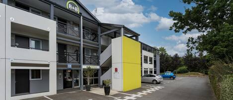 A modern hotel with a yellow accent wall, a parking area with marked spaces, and a car parked in front.