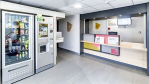 A modern reception area with a vending machine stocked with snacks and drinks, a reception desk with informational posters, and a clean, tiled floor. - B&B HOTEL Chalon-sur-Saône Sud (Saint-Remy)