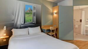 A bedroom with a large bed, a desk, and a view of a mountainous landscape through an open window. - B&B HOTEL Chalon-sur-Saône Sud (Saint-Remy)