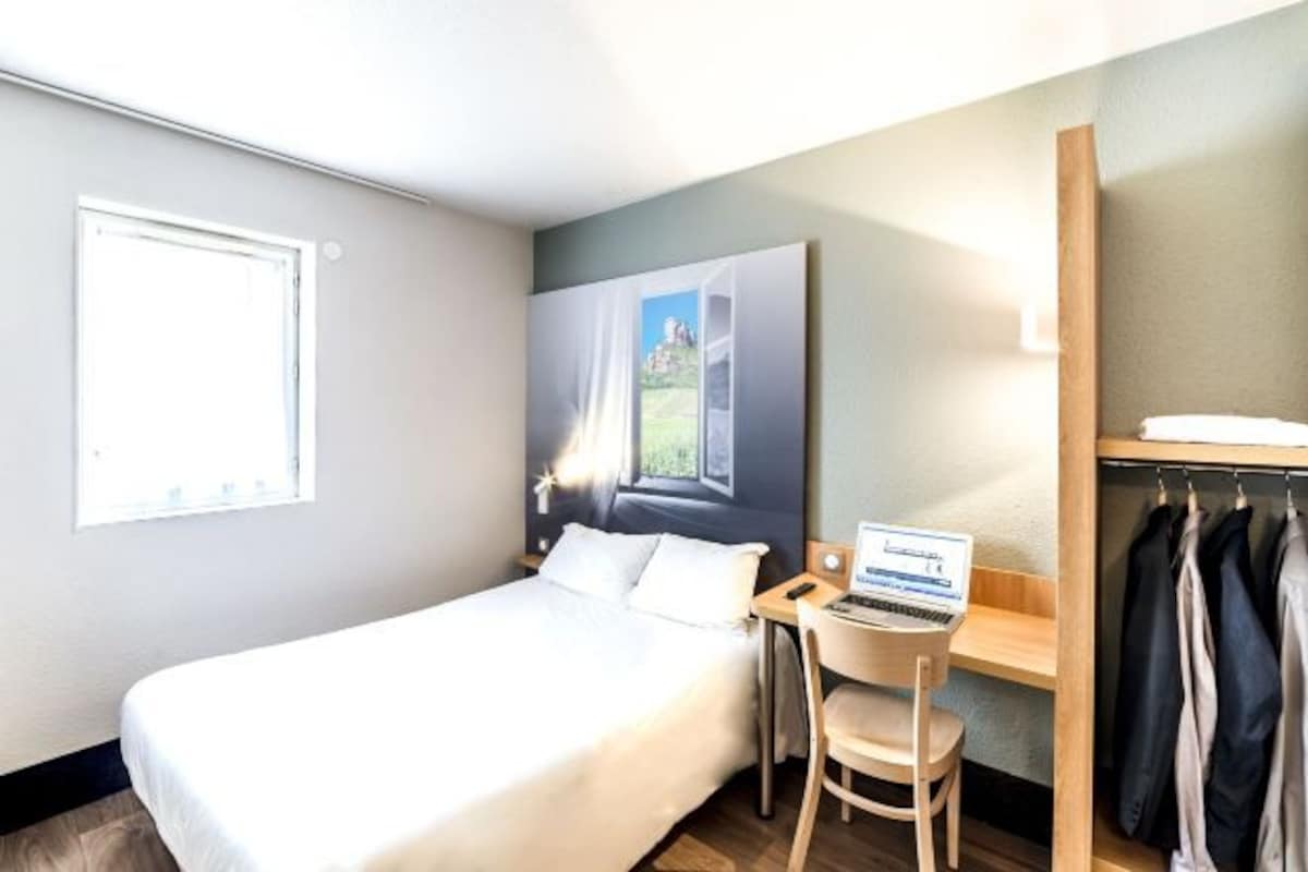 Ein Hotelzimmer mit Bett, Schreibtisch mit Laptop, Stuhl, Kleiderhaken mit Kleidung und einem Fenster.
