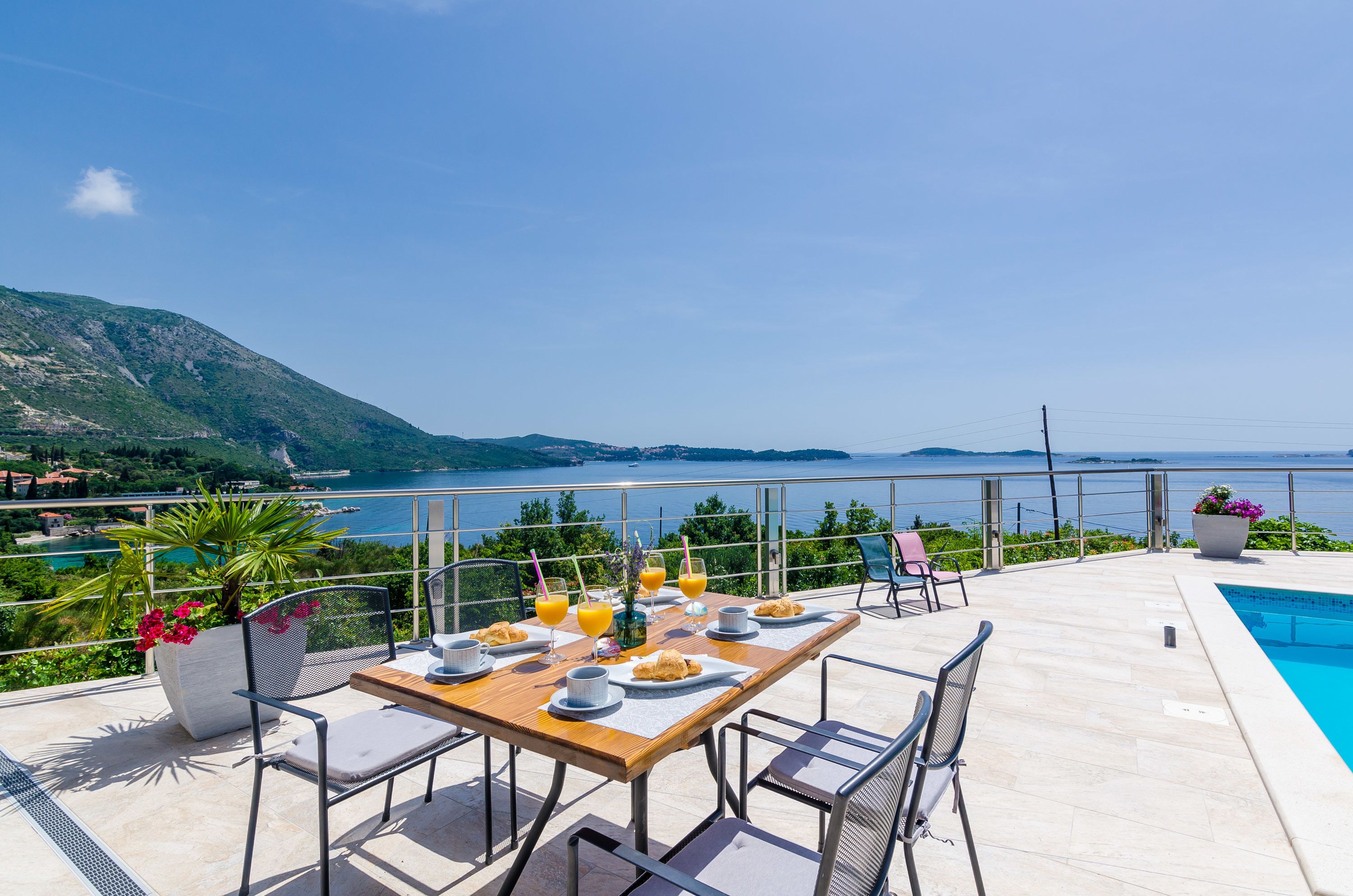 A terrace with a dining table set for two, overlooking a pool and a scenic view of the sea and hills.