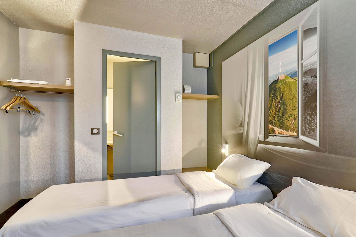 Ein Hotelzimmer mit zwei Betten, einem Spiegel, einem Fenster mit Aussicht und einer Tür zu einem anderen Raum.