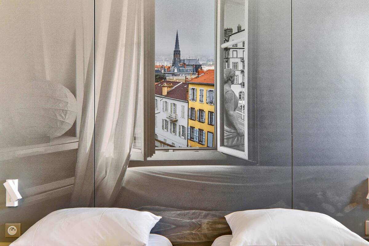 Ein Zimmer mit einem Bett, weißen Kissen, einem Fenster und Blick auf Gebäude sowie einen Kirchturm.