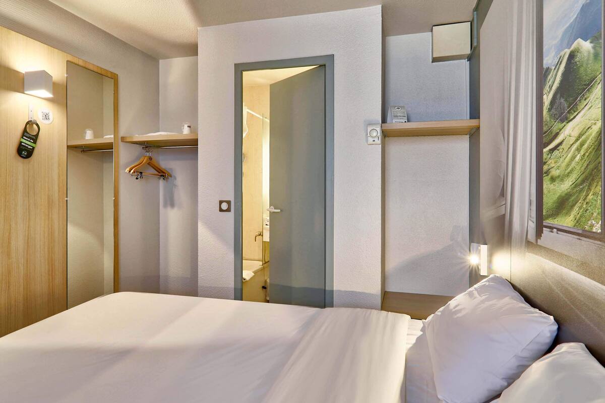 Ein Hotelzimmer mit einem Bett, einem Holzschrank, einem Spiegel, einem Kleiderhaken und einem Landschaftsbild.