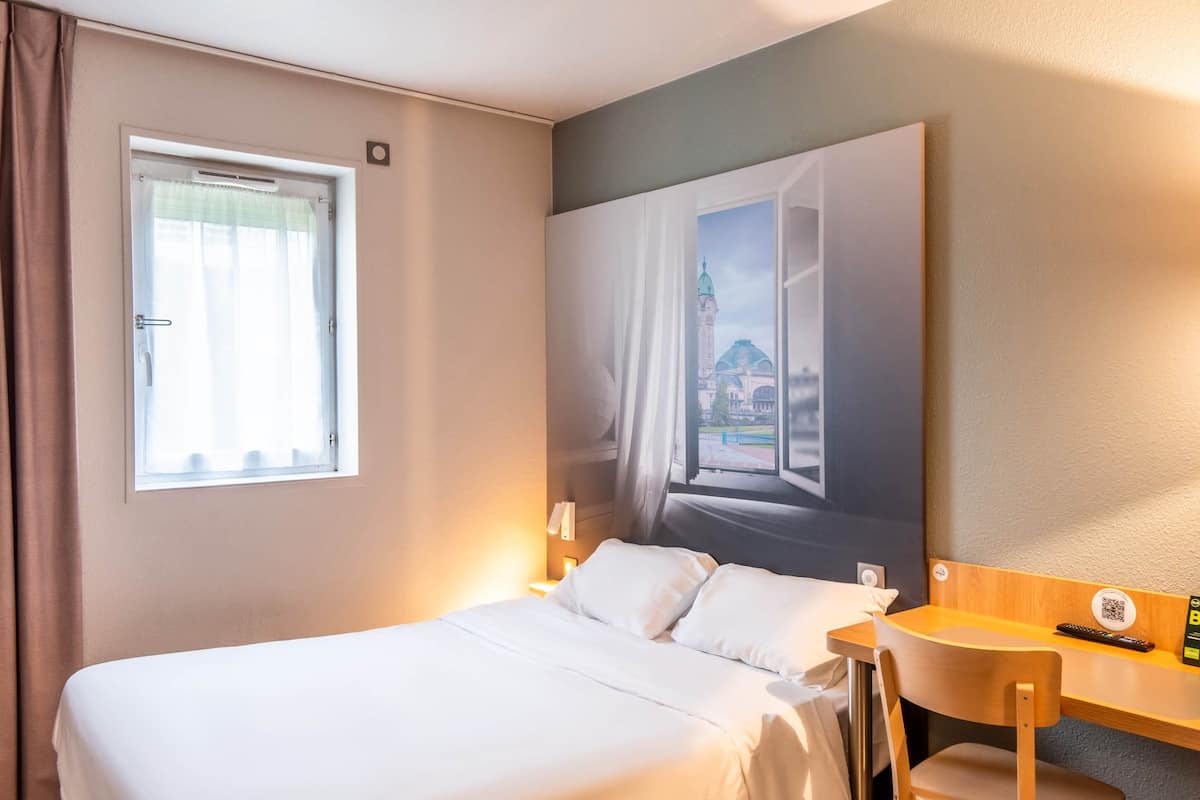 A hotel room with a bed, a desk with a chair, a window with curtains, and a wall-mounted painting.