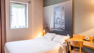 Een hotelkamer met een bed, een bureau met stoel, een raam met gordijnen en een aan de muur hangend schilderij.