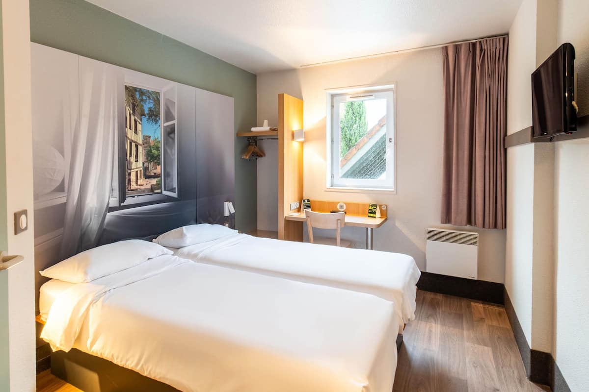 A hotel room with two beds, a desk, a chair, a TV, and a window with curtains.