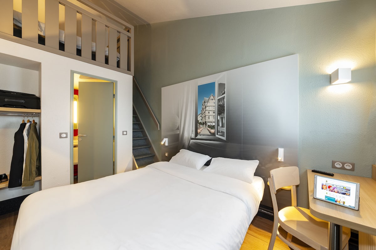 Une chambre d’hôtel comprenant un lit, un bureau avec une tablette, une chaise, un porte-manteau et un escalier.