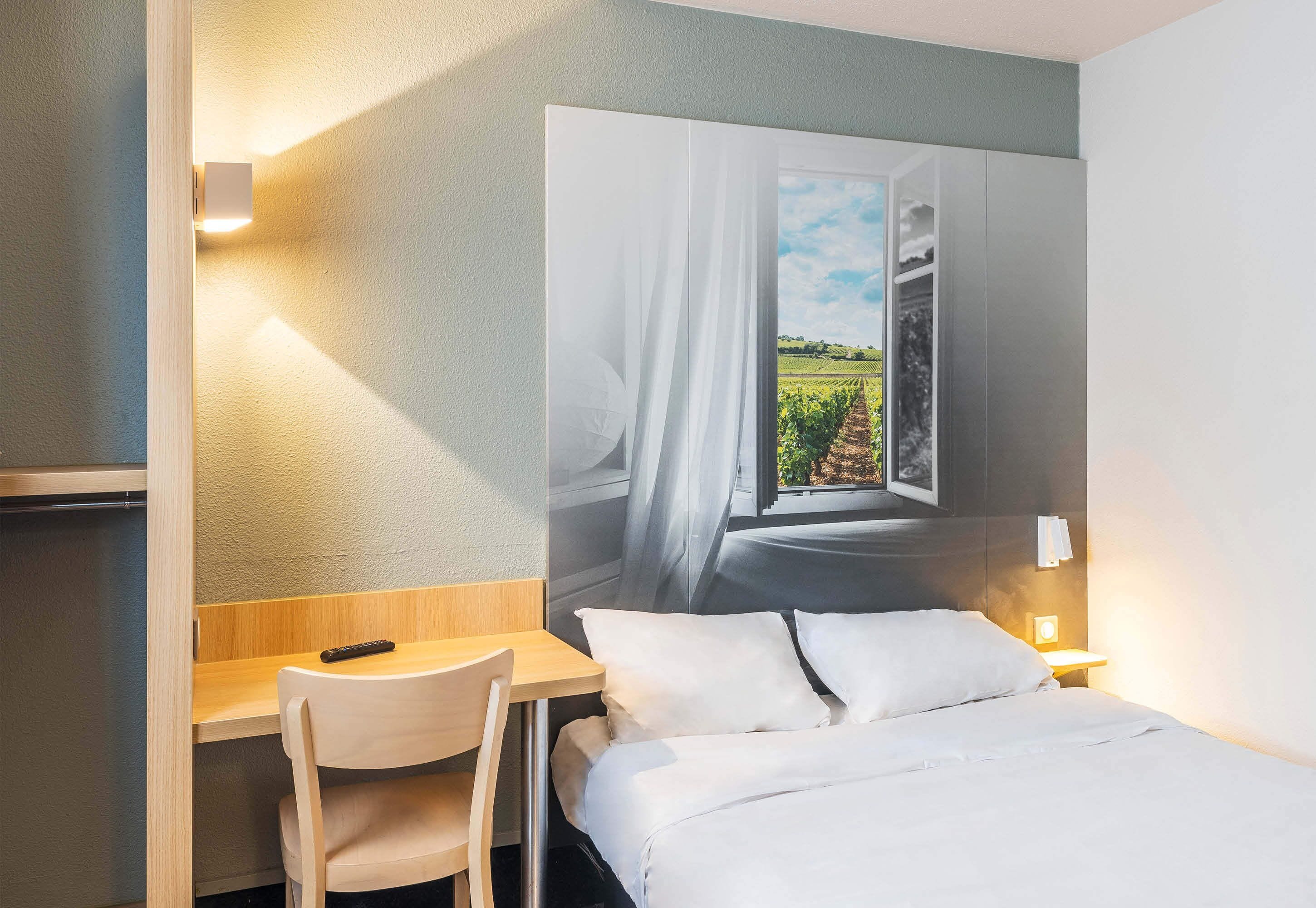 Ett hotellrum med en säng, ett skrivbord, en stol och en väggmonterad TV som visar en utsikt över en vingård.