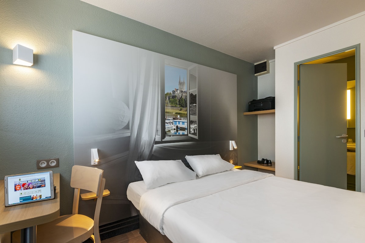 Une chambre d’hôtel moderne avec un grand lit, un bureau avec un ordinateur portable, une chaise et une vue sur le paysage urbain par la fenêtre.