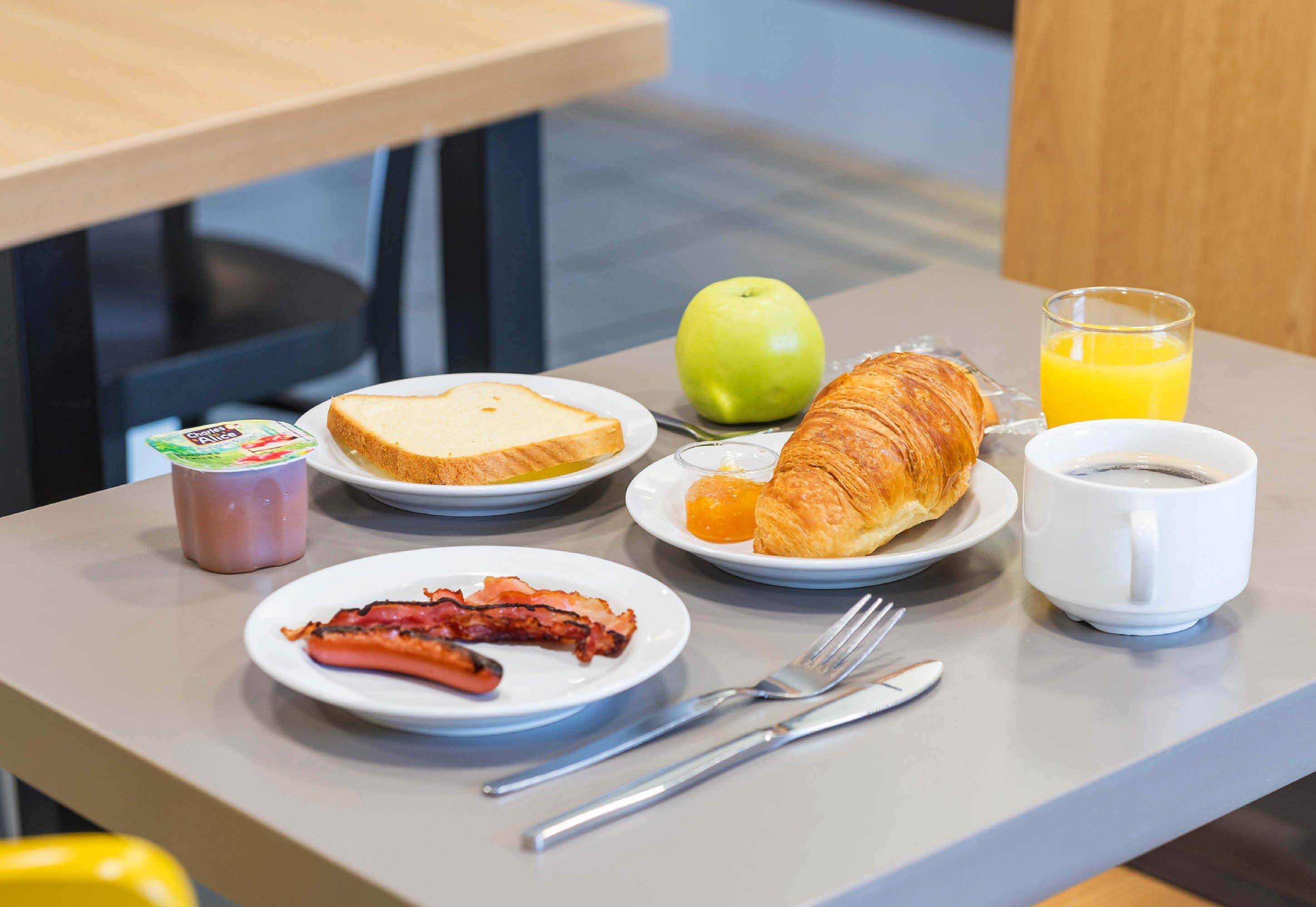 Una tavola imbandita con pane tostato, un croissant, pancetta, una mela, un bicchiere di succo d'arancia, una tazza di caffè e un vasetto di yogurt.