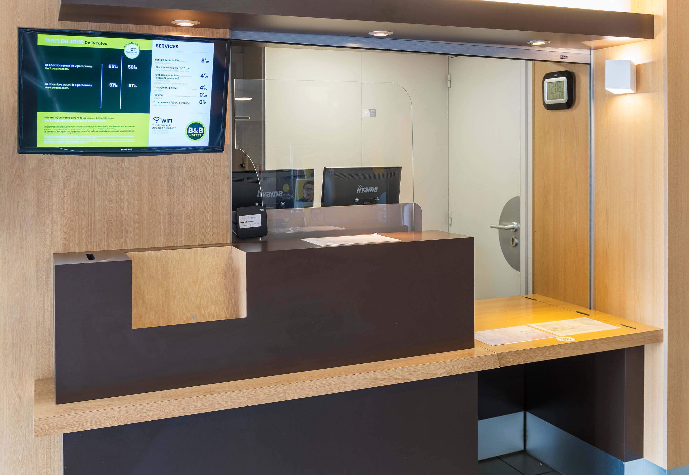A reception desk with a digital display, a wooden counter, and a glass partition.