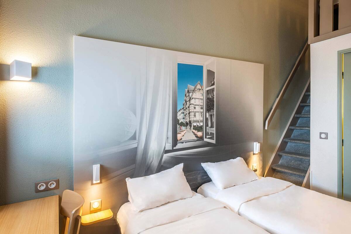 Une chambre d’hôtel avec un lit, des tables de chevet, une chaise, un tableau représentant un bâtiment et un escalier.