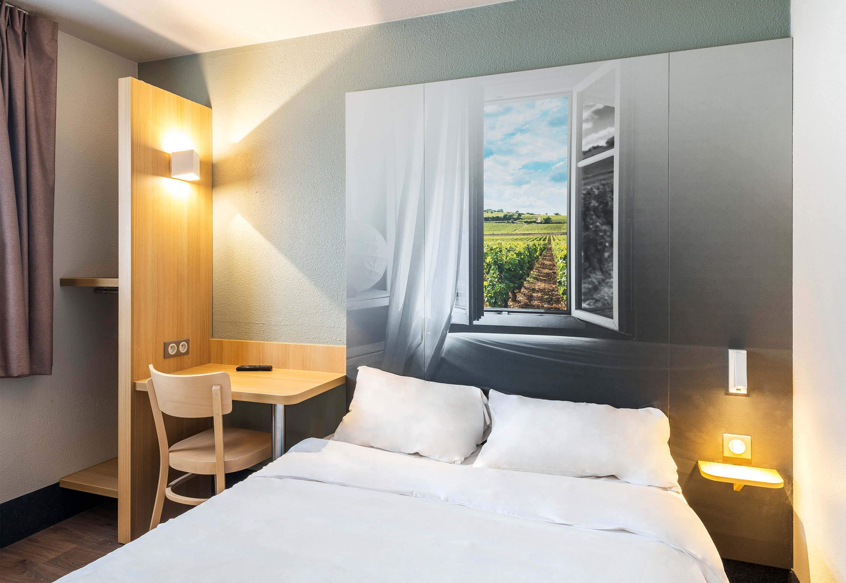 A hotel room with a bed, a desk, and a chair. There is a window with a view of a vineyard.