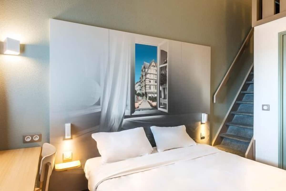 Une chambre d’hôtel avec un lit, une table de chevet, une lampe fixée au mur, un escalier et une grande fenêtre donnant sur des immeubles.