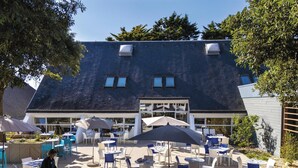 An outdoor patio area with tables and chairs, a person arranging items, and a building with a sloped roof in the background. - Belambra Clubs Guidel - Les Portes De L'Océan (Guidel)