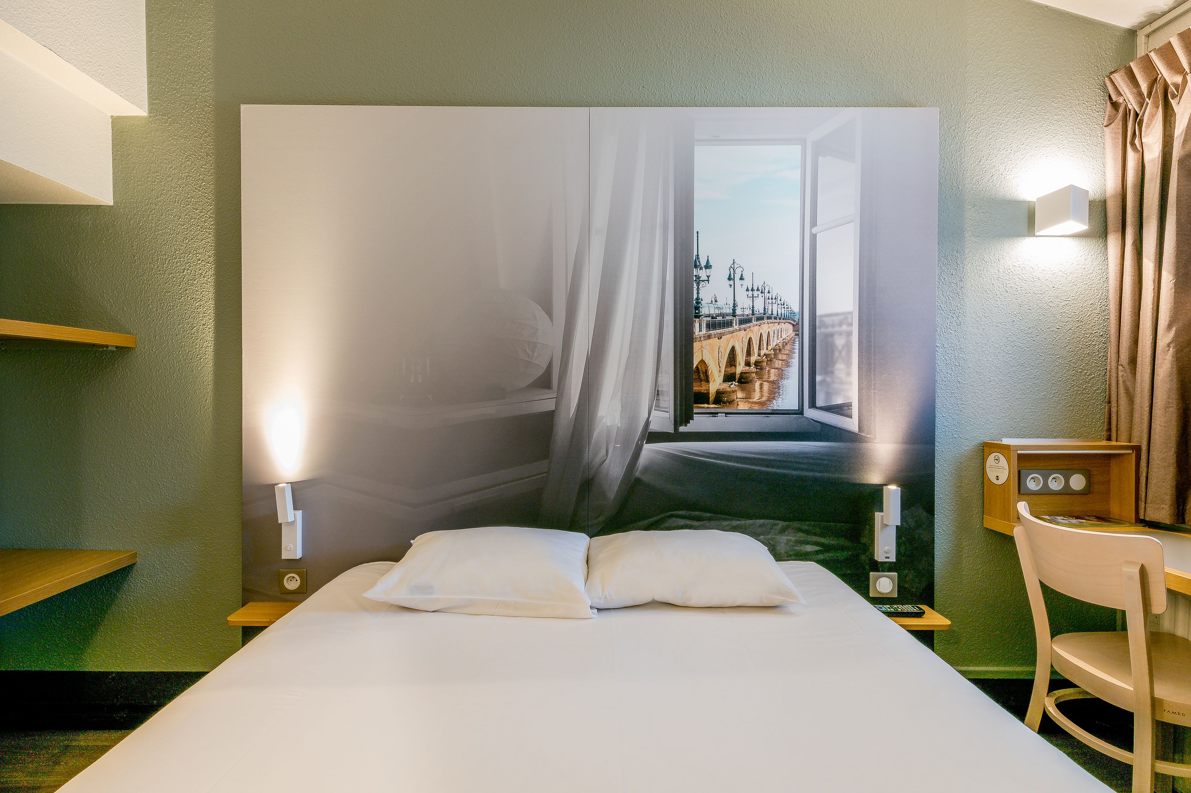 A hotel room with a large bed, a desk, a chair, a nightstand, and a wall-mounted lamp.
