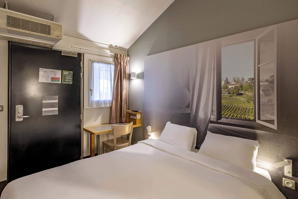 Quarto de hotel com cama, escrivaninha, cadeira, aparelho de ar condicionado, janela com cortinas e luminária na parede.
