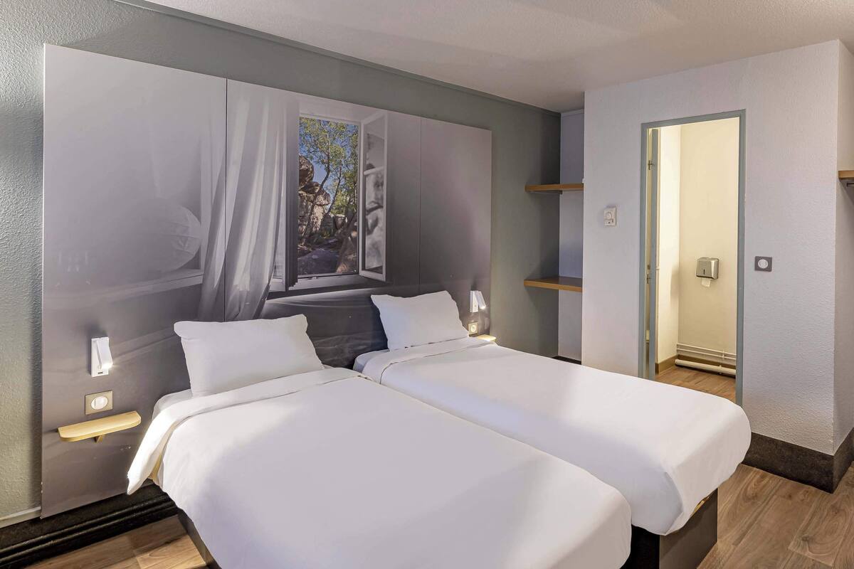 Quarto de hotel com duas camas, uma janela grande com vista, uma mesa de cabeceira e uma porta que dá para outro cômodo.