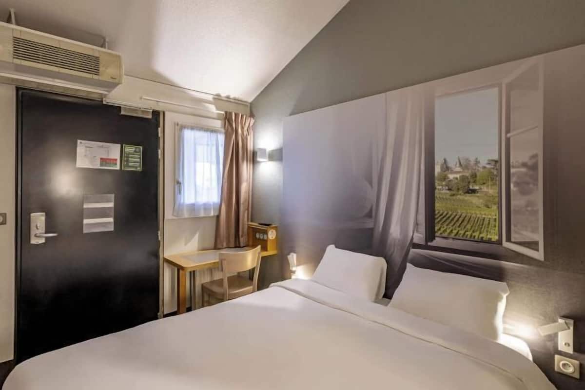 A hotel room with a bed, a desk, a chair, a window with curtains, and an air conditioning unit.