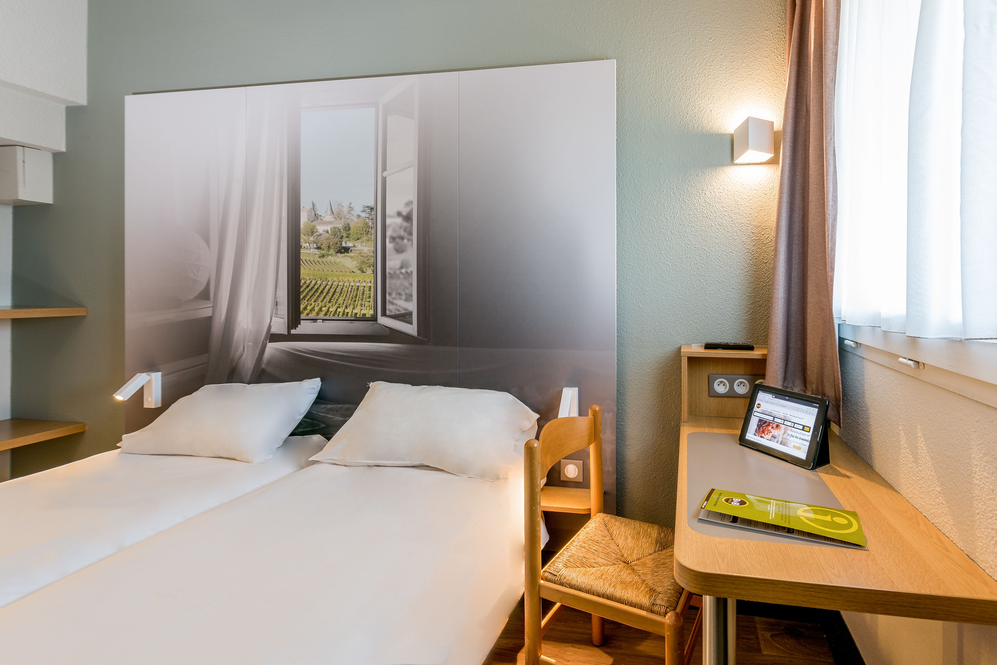 A hotel room with a bed, a desk with a tablet, a chair, and a large wall mural.
