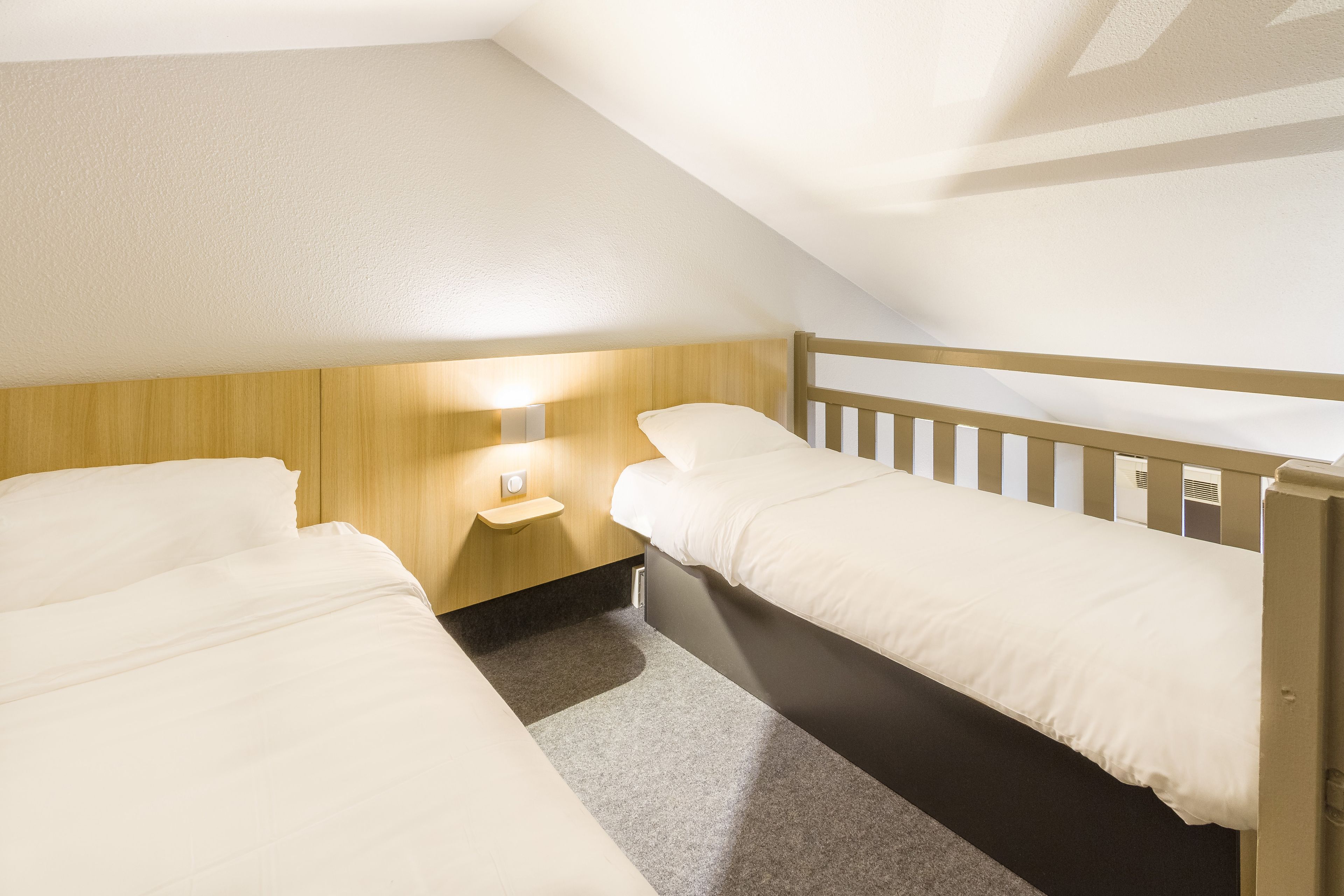 Una habitación pequeña y luminosa con dos camas individuales, una mesita de noche con lámpara y un cabecero de madera.