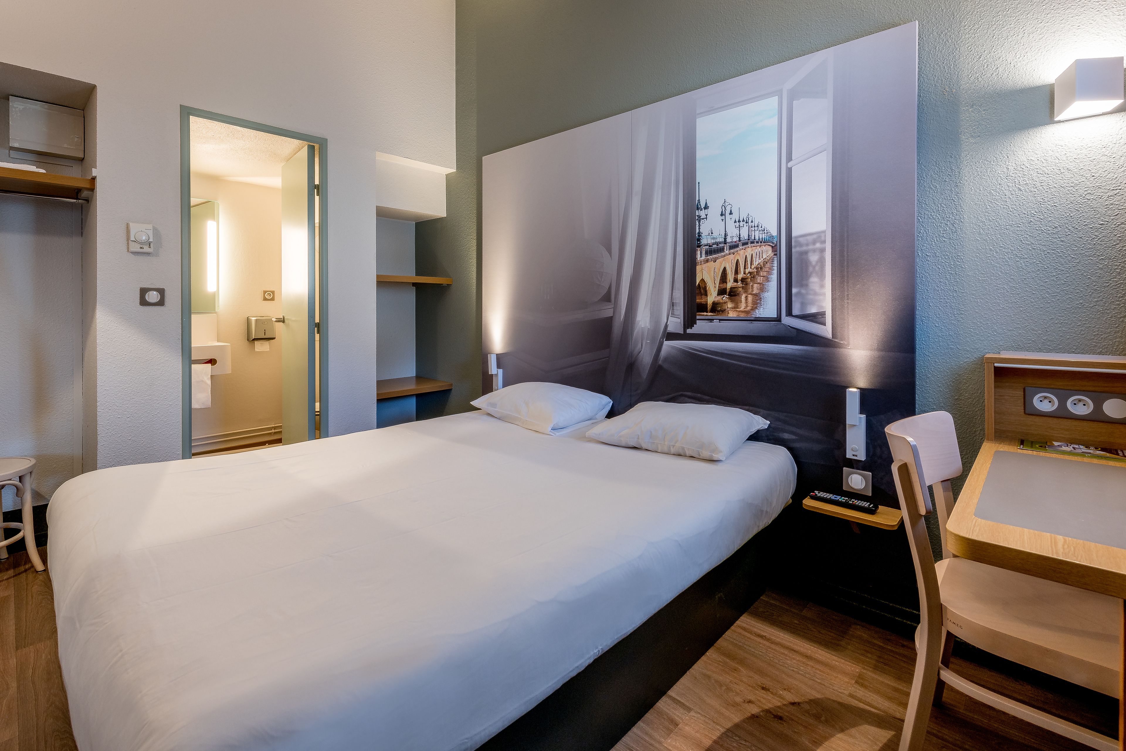 A hotel room with a large bed, a desk, and a chair. There is a window with a view of a bridge.