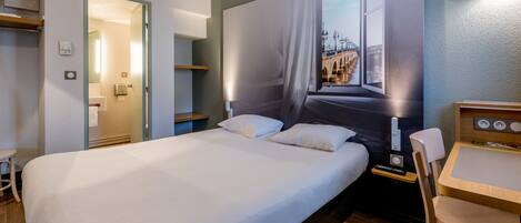A hotel room with a large bed, a desk, and a chair. There is a window with a view of a bridge.