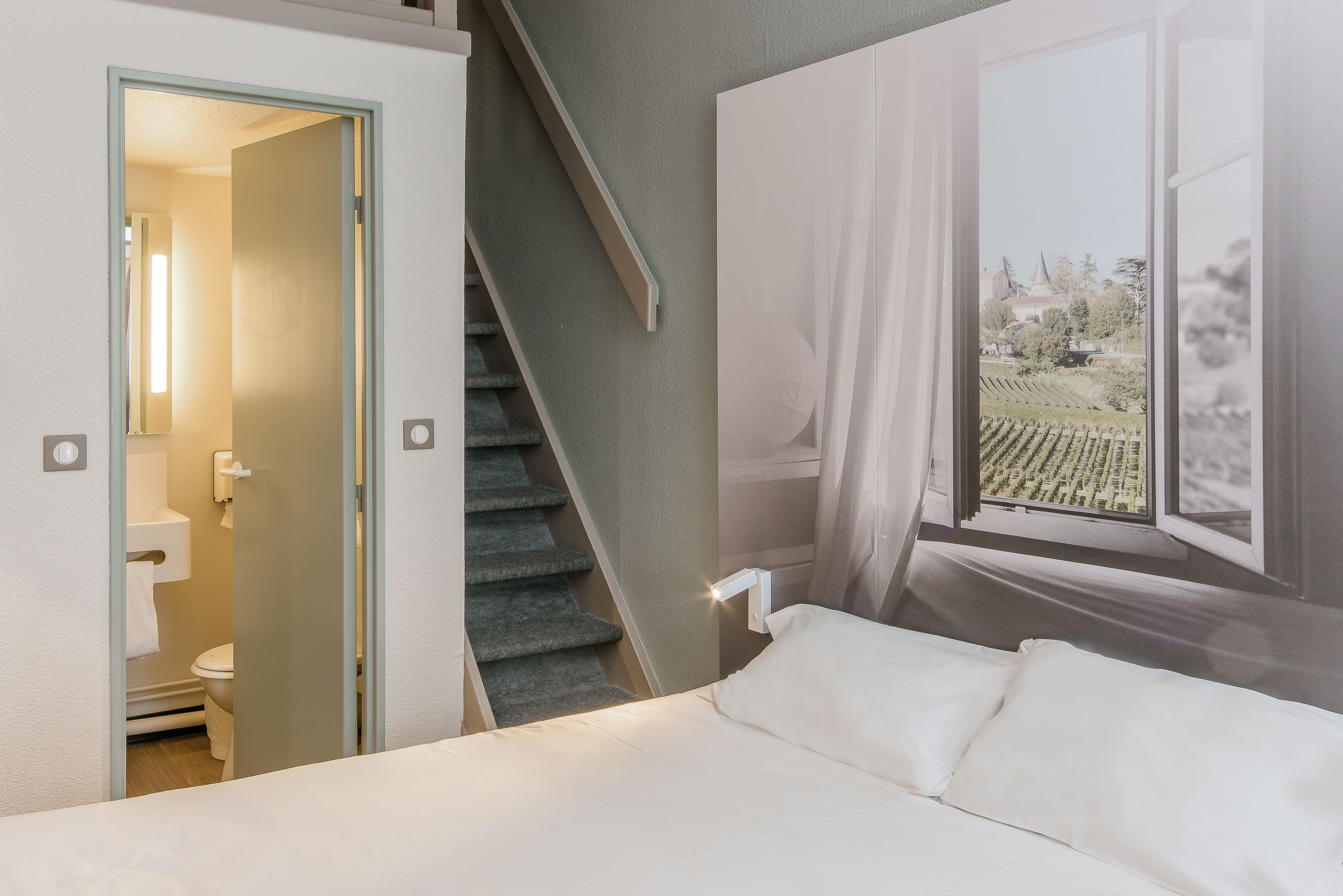 Un dormitorio con cama, ventana con vistas a viñedos, escalera y baño visible a través de una puerta abierta.