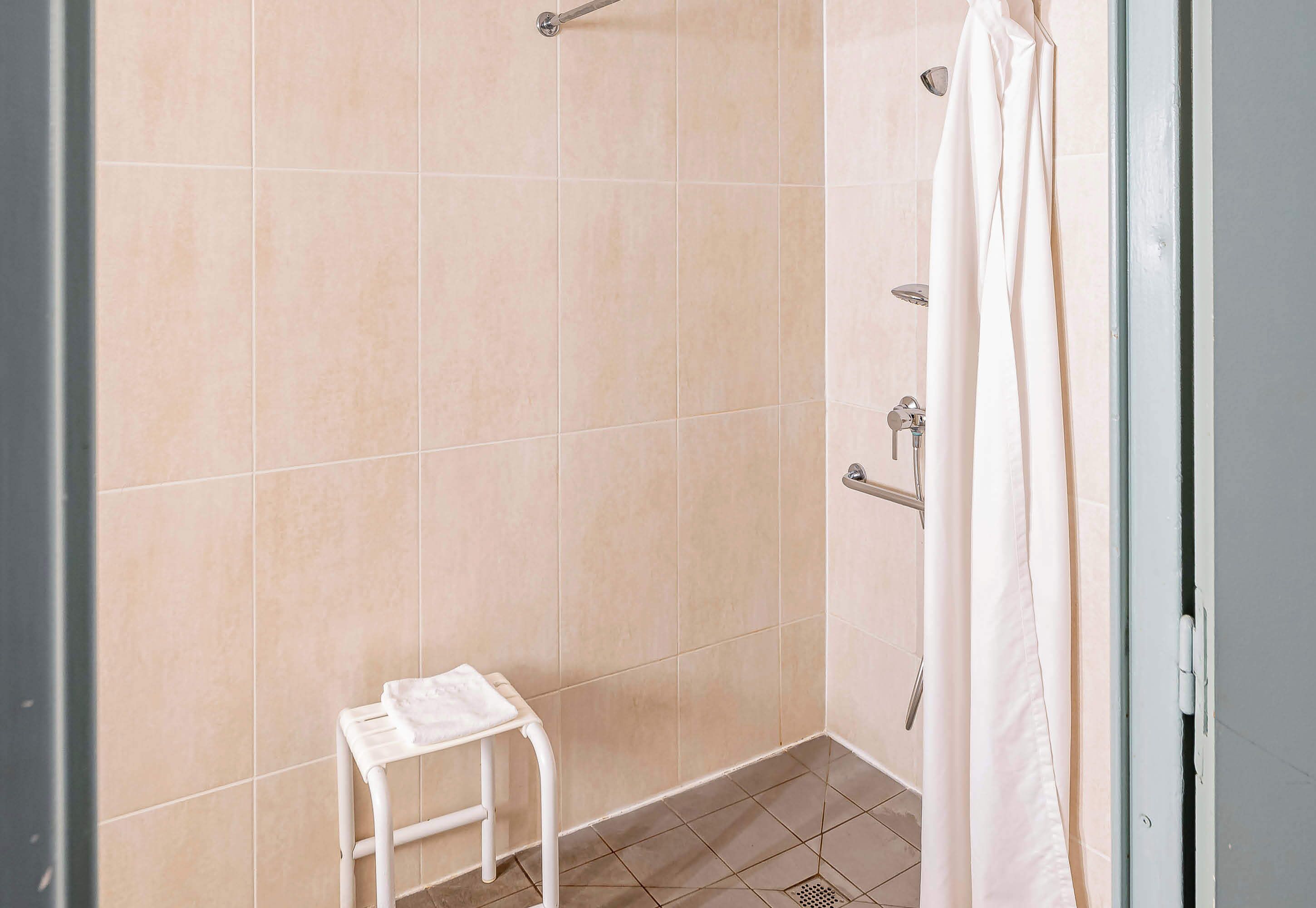 Un espacio de ducha con azulejos color beige, una toalla blanca sobre un taburete pequeño y una cortina de ducha blanca.