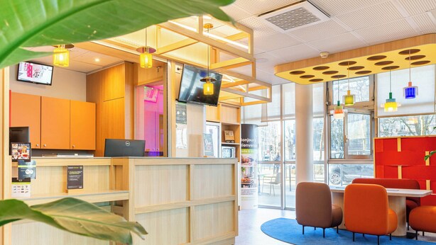 A reception area with a wooden desk, a computer, and a TV screen. There are chairs and a table in the background. - B&B Hôtel Nantes Centre (Nantes)
