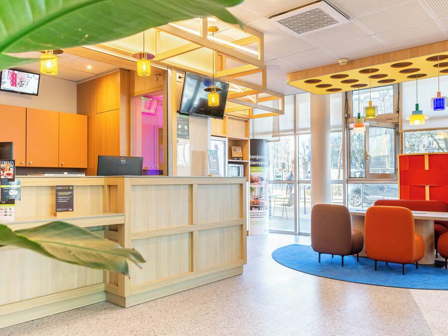 A reception area with a wooden desk, a computer, and a TV screen. There are chairs and a table in the background.