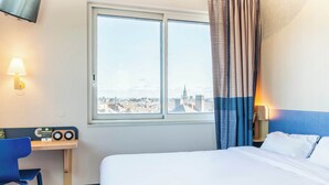 A hotel room with a bed, a desk with a radio, a chair, a window with a city view, and a TV mounted on the wall. - B&B Hôtel Nantes Centre (Nantes)