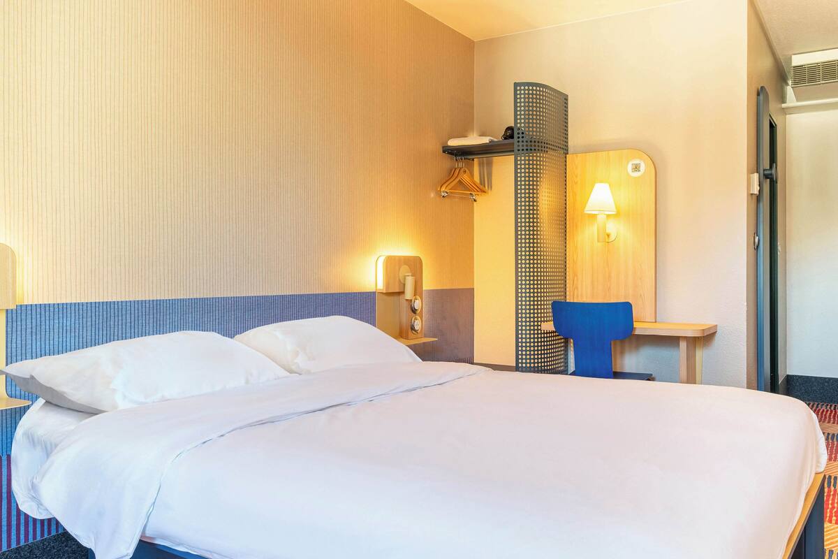 Habitación de hotel con una cama grande, un escritorio con silla, una lámpara de pared y un perchero.