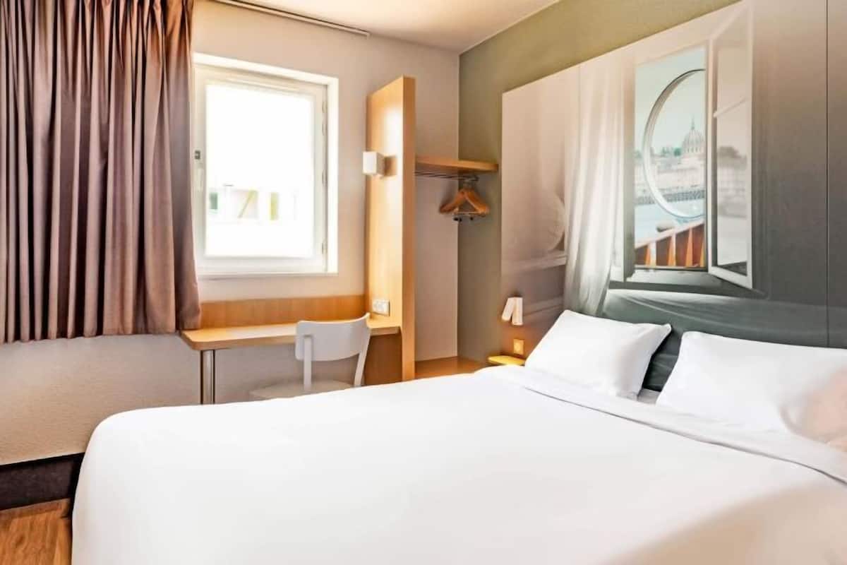 Ein Hotelzimmer mit Bett, Schreibtisch, Stuhl, einem Fenster mit Vorhängen und Blick auf ein Gebäude.