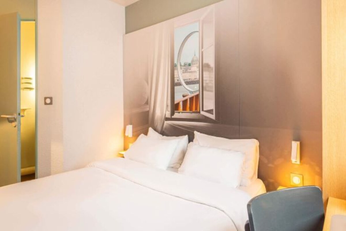 Een hotelkamer met een bed, een stoel, een raam met gordijnen en een wandlamp.