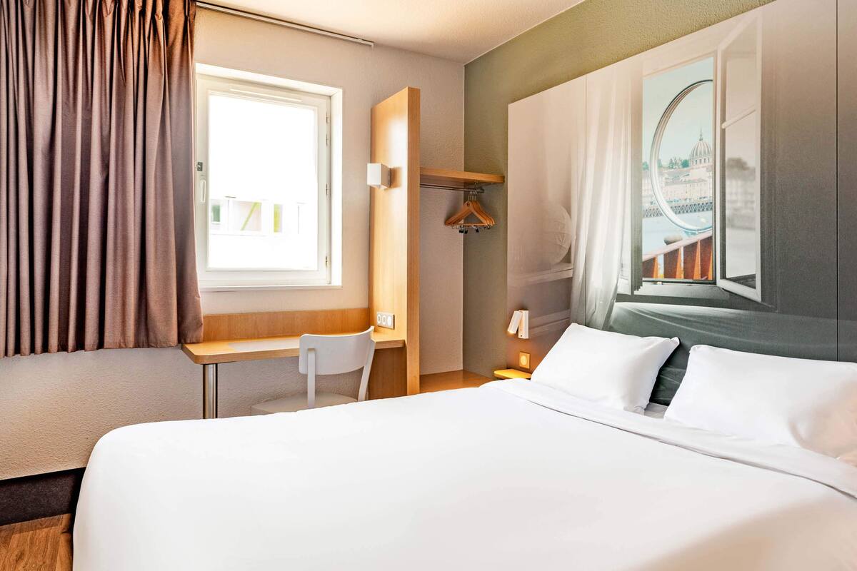 Ein Hotelzimmer mit einem Bett, einem Schreibtisch, einem Stuhl, einem Kleiderständer und einem Fenster mit Blick auf eine Brücke.