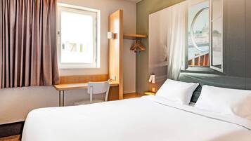 A hotel room with a bed, a desk, a chair, a coat rack, and a window with a view of a bridge.