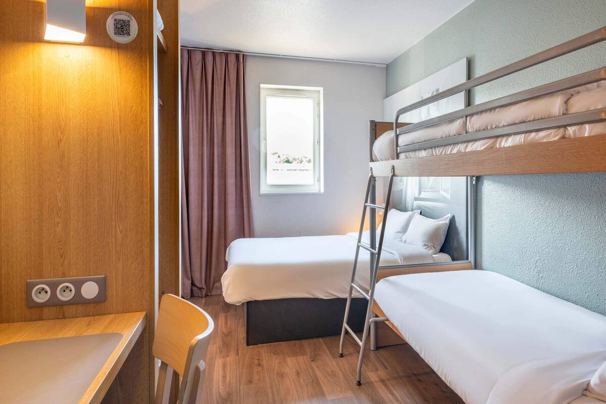 Ein kleines Hotelzimmer mit Etagenbetten, einem Schreibtisch, einem Stuhl, einem Fenster mit Vorhängen und einem QR-Code an der Wand.